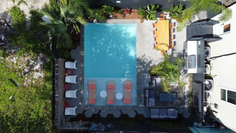 Aerial drone view of a turquoise rectangular backyard pool with three submerged lounge chairs, a row of sunbeds, palm trees, patio seating and a wooden outdoor dining table.