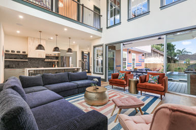 Modern open-concept living room with double-height ceiling, large blue sectional, rust-orange accent chairs, patterned area rug and brass coffee table, kitchen island with pendant lights, and sliding glass doors opening to a backyard patio and pool.