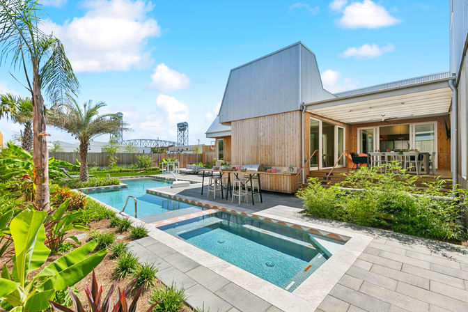 Sunlit backyard with modern wood-clad house, rectangular pool and built-in spa, palm trees, lush tropical landscaping, and covered outdoor dining patio