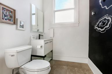 Bright minimalist bathroom with white toilet and floating vanity, black faucet and rectangular mirror, sunlit window, black accent wall with white abstract design, framed artwork and beige tile floor.