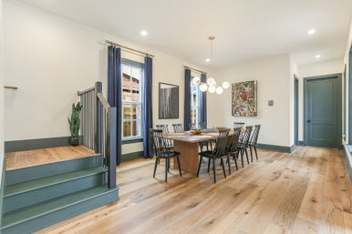 Bright modern home dining room interior with long wood table and eight black spindle chairs, light oak hardwood floors, globe pendant chandelier, navy-trim windows with curtains, small staircase landing and potted plant, minimalist wall art.