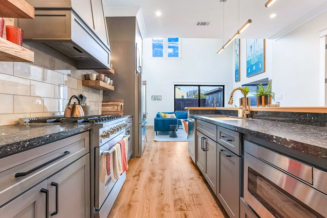 Bright, airy modern open-concept kitchen with gray cabinets, black speckled countertops, stainless steel range and appliances, light hardwood floors, brass sink faucet, pendant lighting, open shelves, and a blue-sit living area visible through a sliding glass door.
