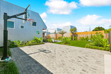 Sunny modern backyard with paver basketball half-court and hoop, corrugated metal house, green lawn, palm trees and patio seating