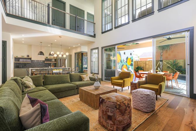 Bright two-story open-plan living room with a large green sectional, mustard armchairs, wooden coffee table and patterned poufs, modern kitchen with pendant lights, and sliding glass doors opening to a colorful patio mural and outdoor dining area.