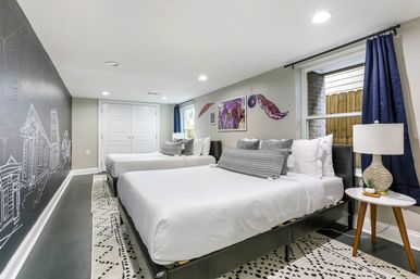 Bright modern guest bedroom with two beds in white linens, decorative gray pillows, chalkboard wall mural of a city skyline, patterned rug, navy curtains, bedside table with lamp and window — stylish urban vibe.