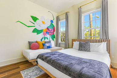 Sunlit modern bedroom with two beds, a bold colorful floral wall mural, wooden slat headboard, gray curtains, decorative throw pillows and warm hardwood floors