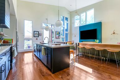 Sunlit modern open-concept kitchen and living area featuring a dark central island with quartz countertop, globe pendant lights, hardwood floors, vaulted ceilings, teal TV accent wall and wooden breakfast bar with stools.