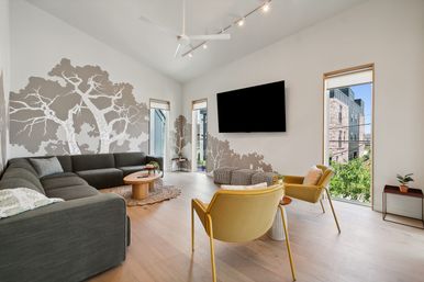 Bright modern urban living room in condo with vaulted ceiling and fan, gray sectional sofa, tree-silhouette wall mural, wall-mounted TV, light wood floors, mustard accent chairs, round wooden coffee table and tall narrow windows with city view.