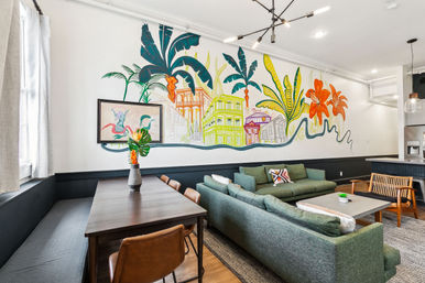 Open-plan modern loft living and dining area with a large colorful tropical mural of palm leaves and pastel houses, green sectional sofa, wooden dining table with leather chairs, pendant lighting and a vase of tropical flowers.