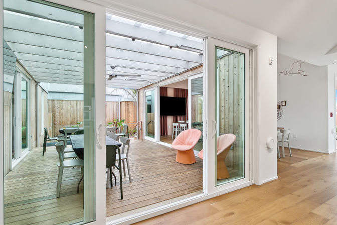 Bright indoor-outdoor living space with large sliding glass doors leading to a covered wooden deck patio, modern dining table and chairs, two coral-pink lounge chairs, ceiling fan and wall-mounted TV.