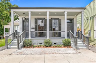 Symmetrical gray duplex with covered porch, four white columns, twin staircases and black shutters, small gravel planting bed and shrubs under a bright blue sky