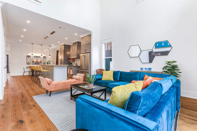 Open-concept modern living room and kitchen with a vibrant teal velvet sectional, colorful throw pillows, pink chaise lounge, hardwood floors, black coffee table, hexagonal wall mirrors, and a kitchen island lined with yellow bar stools and stainless-steel appliances.