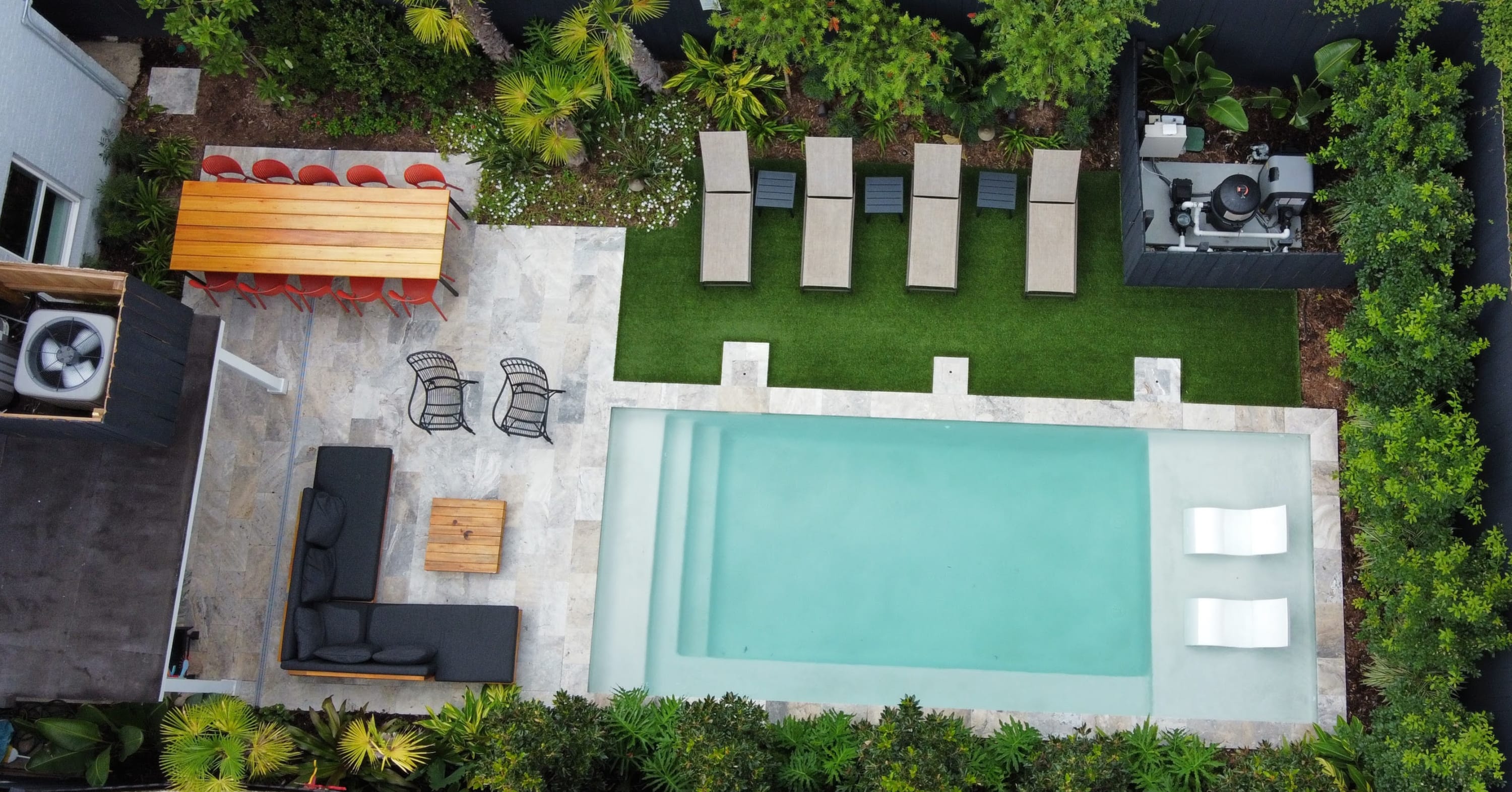 Aerial drone view of a modern residential backyard: rectangular turquoise pool with two submerged white loungers, stone patio with dark sectional sofa and wooden coffee table, long wooden dining table with red chairs, grassy strip with four tan sunbeds, and lush tropical landscaping.