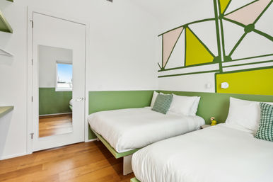Bright modern twin bedroom with two white-dressed beds, green half-wall and geometric mural, hardwood floors, full-length mirror on the door, green patterned throw pillows and small yellow bedside lamp.