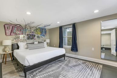 Modern bedroom with king bed, colorful hand-drawn city mural above the headboard, navy curtains, full-length mirror and patterned gray area rug