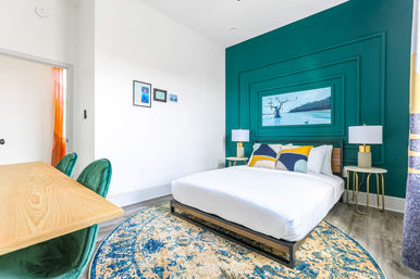 Bright modern bedroom with teal feature wall, white bed with colorful pillows, round blue-beige patterned rug, wooden platform frame, gold-accent bedside tables and lamps, framed artwork above bed, wooden desk and green velvet chairs—contemporary apartment interior.