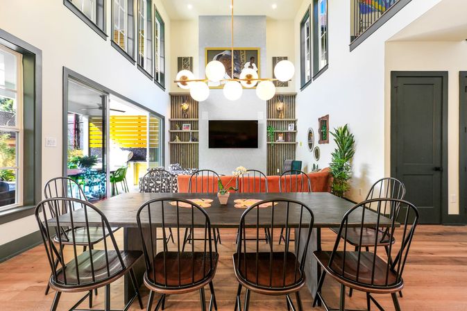 Bright modern two-story open-concept dining and living room with a long dark wood table and black metal chairs, orange sofa, wall-mounted TV, globe pendant chandelier, sliding glass doors to patio, hardwood floors and plants