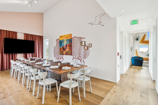 Beach-themed communal dining room with a long wooden table set for service, white chairs, ocean and seagull wall murals, large wall-mounted TV, pink curtains and a hallway leading to blue lounge chairs and coastal murals.