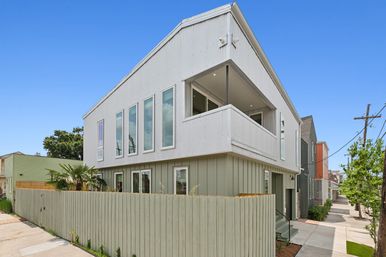 Contemporary two-story coastal-style house on a corner lot with tall vertical windows, covered second-floor balcony, pale siding, fenced yard and clear blue sky