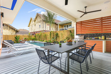 Sunlit modern covered backyard patio with outdoor dining table for six, woven chairs, gas grill and bar, lounge chairs beside a rectangular pool, palm plants and wooden privacy fence.