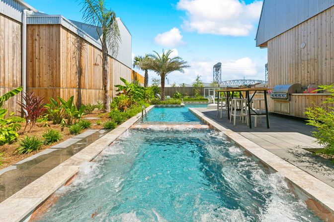Sunny modern backyard with a long lap-style pool and bubbling spa in the foreground, palm tree and tropical landscaping, patio bar with stools and built-in grill, and wooden privacy fences.