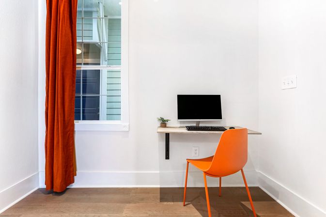 Minimal modern home office nook in a white-walled room with a wall-mounted desk holding a black monitor and keyboard, small potted plant, a cheerful orange chair, and matching orange curtain by the window.