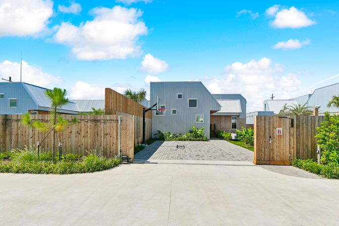Sunny modern coastal home with metal siding, wooden privacy fence and gate, paved driveway with basketball hoop and tropical landscaping under a blue sky