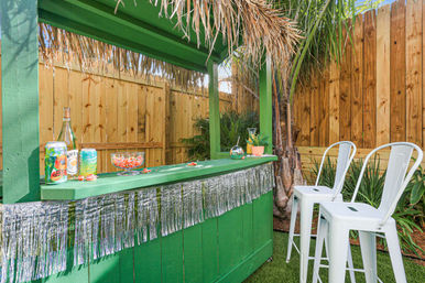 Sunny backyard tropical-themed green tiki bar with thatched roof and silver fringe, drinks and a candy bowl on the counter, two white metal bar stools, palm plants and wooden fence backdrop.