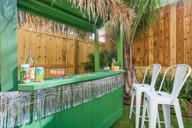 Sunny backyard tropical-themed green tiki bar with thatched roof and silver fringe, drinks and a candy bowl on the counter, two white metal bar stools, palm plants and wooden fence backdrop.