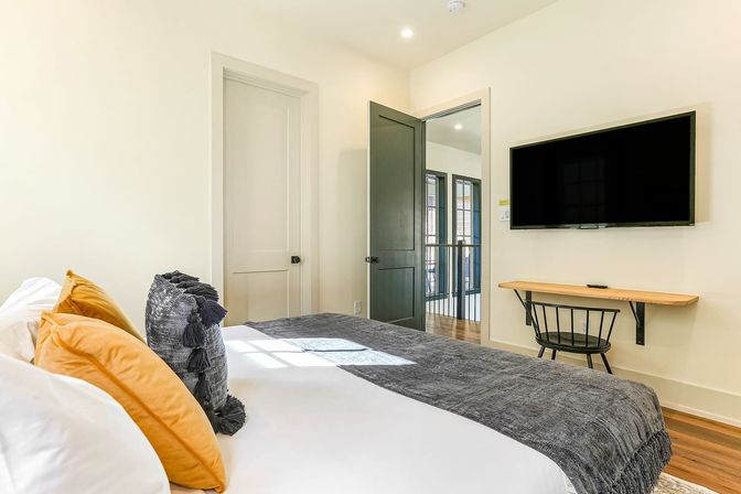 Cozy bright contemporary bedroom interior with white walls, bed styled with mustard and charcoal pillows and a gray throw, wall-mounted TV above a floating wooden desk and black chair, open dark-gray door and warm hardwood floors.