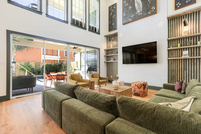 Sunlit double-height living room with olive-green sectional sofa, wall-mounted TV, wooden coffee table, built-in vertical-slat shelving, and wide sliding glass doors opening to a covered patio with outdoor dining and pool view.