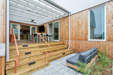 Modern wooden backyard patio with covered deck, orange handrails, outdoor dining table and chairs, wall-mounted TV and ceiling fan, and a gray cushioned lounge on stone pavers against vertical cedar siding.