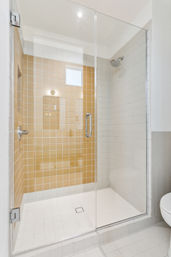 Bright modern walk-in shower with frameless glass door, beige accent tiles on back wall, white tiled floor and walls, chrome rain showerhead and handle in a contemporary residential bathroom