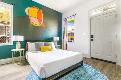 Bright modern bedroom with abstract mural on teal accent wall, white bed with yellow throw pillow, round blue rug, gold bedside tables with lamps, patterned curtains and a front door