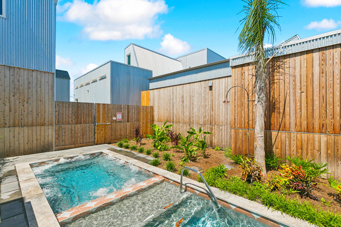 Sunny townhouse courtyard with bubbling hot tub spa, metal handrail, outdoor shower, palm tree, landscaped tropical plants and wood privacy fence against modern metal-clad buildings.