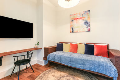 Cozy modern sitting area with a wooden daybed draped in a blue throw and colorful pillows, wall-mounted TV, floating wood desk, abstract wall art and warm hardwood floors