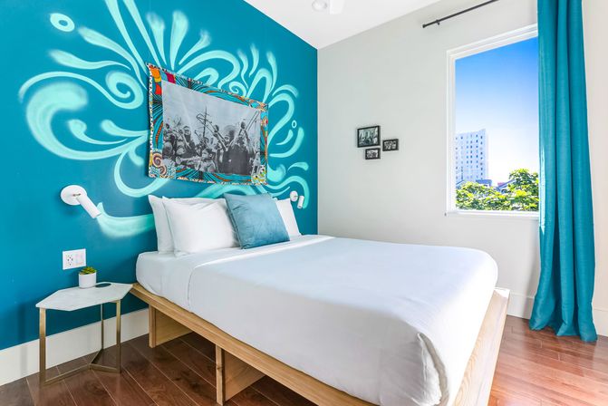 Bright modern bedroom with white linens on a wooden platform bed, turquoise accent wall featuring a swirling mural and framed black-and-white artwork, teal curtain framing a sunny city-view window and a small marble side table with a potted plant.