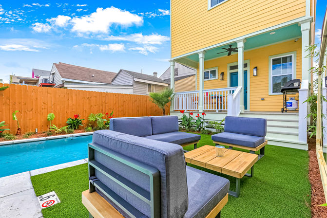 Backyard of a yellow two-story house with covered porch and teal door, blue outdoor sofa set and wooden coffee table on green turf beside a rectangular pool, wooden fence and grill under a bright blue sky