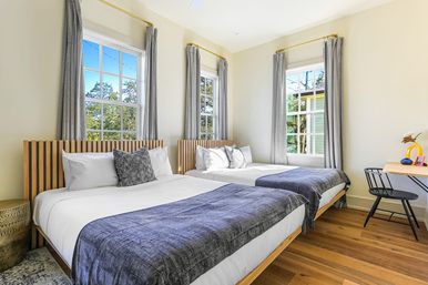 Sunlit modern guest bedroom with two beds featuring slatted wood headboards, crisp white linens and gray throws, three tall windows with gray curtains, hardwood floors and a small desk and chair.