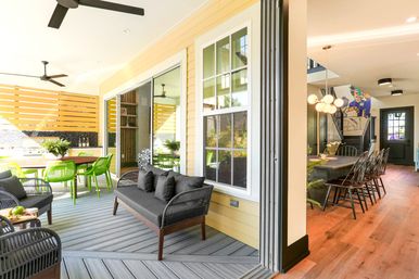 Bright covered patio and open-plan indoor-outdoor living area with yellow siding, gray composite deck, black wicker sofa, lime-green dining chairs, ceiling fans and sliding glass doors to a dining room with pendant lights.