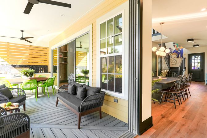 Bright covered patio and open-plan indoor-outdoor living area with yellow siding, gray composite deck, black wicker sofa, lime-green dining chairs, ceiling fans and sliding glass doors to a dining room with pendant lights.