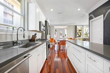 Bright open-concept modern kitchen with white cabinetry, gray quartz counters, stainless steel appliances and sink, long island, polished hardwood floors and orange dining chairs by a glass-paneled front door