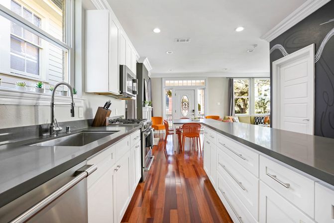 Bright open-concept modern kitchen with white cabinetry, gray quartz counters, stainless steel appliances and sink, long island, polished hardwood floors and orange dining chairs by a glass-paneled front door