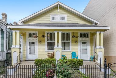 Charming yellow bungalow duplex with a covered front porch, white columns, hanging porch swing, turquoise Adirondack chairs, small landscaped front garden and black wrought-iron fence under a clear blue sky.