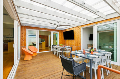 Bright covered wood-deck patio with translucent pergola roof, large outdoor dining table set for eight, orange lounge chairs, wall-mounted TV, ceiling fan and sliding glass doors to a modern kitchen.