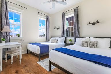 Sunny modern guest bedroom with two white-dressed double beds and blue runners, hardwood floors, large windows with gray curtains, small white desk, ceiling fan and potted plants