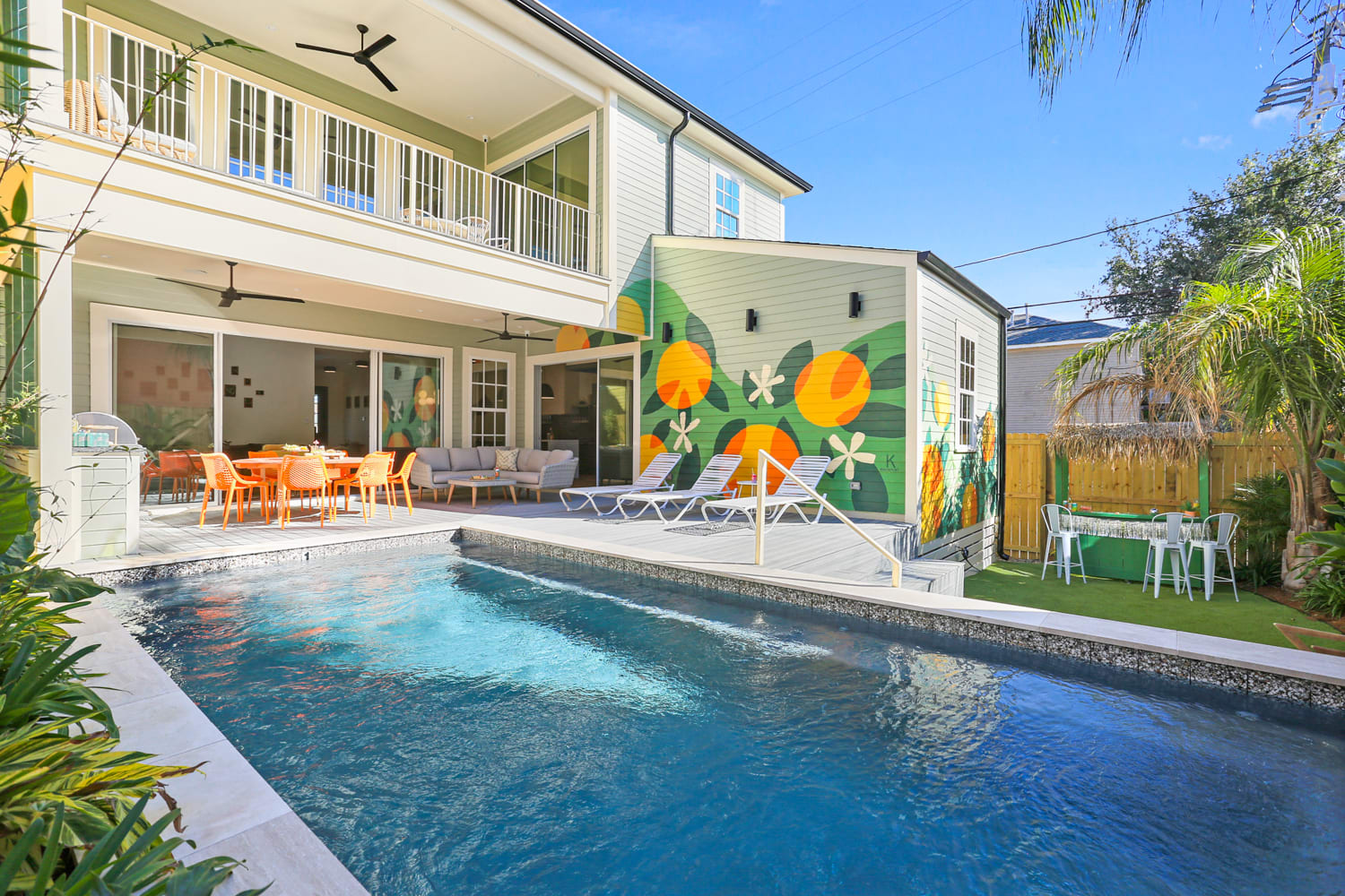 Sunny backyard of a two-story home with a rectangular pool, covered patio with orange outdoor dining set and sofa, deck loungers, vibrant citrus mural and tropical landscaping.