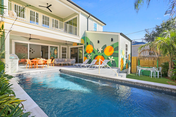 Sunny backyard of a two-story home with a rectangular pool, covered patio with orange outdoor dining set and sofa, deck loungers, vibrant citrus mural and tropical landscaping.