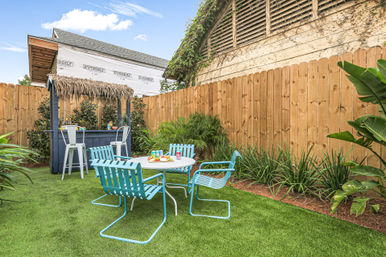 Small fenced backyard patio with tiki-style bar, bright turquoise metal chairs around a white round table on a green lawn, tropical plants and wooden privacy fence