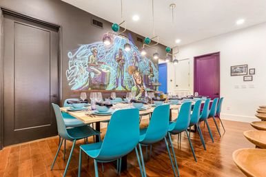 Contemporary dining room interior with a long wooden table set for about twelve, bright turquoise chairs, geometric pendant lights, colorful jazz-themed mural on a dark accent wall, hardwood floors and a purple door.